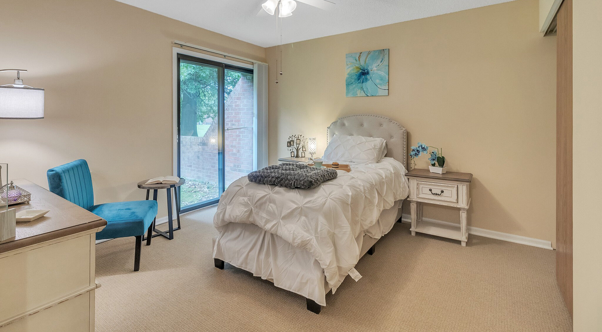 A bedroom with a twin-sized bed with white bedding and a gray throw blanket, a painting of a blue flower above the bed, and a blue chair next to it.