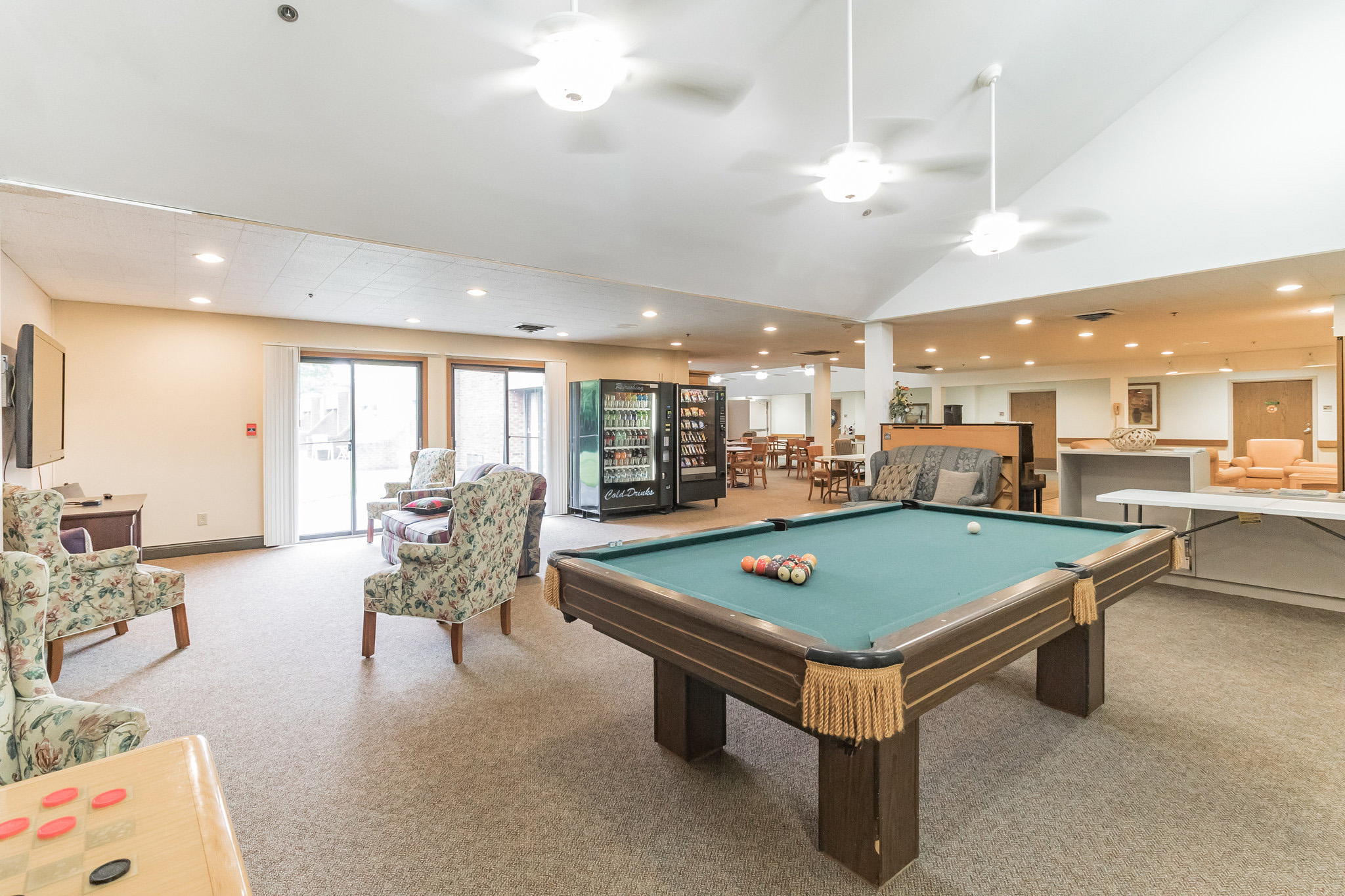 A lounge room with a billiard table in the middle, flower-patterned sofa chairs, a vending machine, and a grey couch.