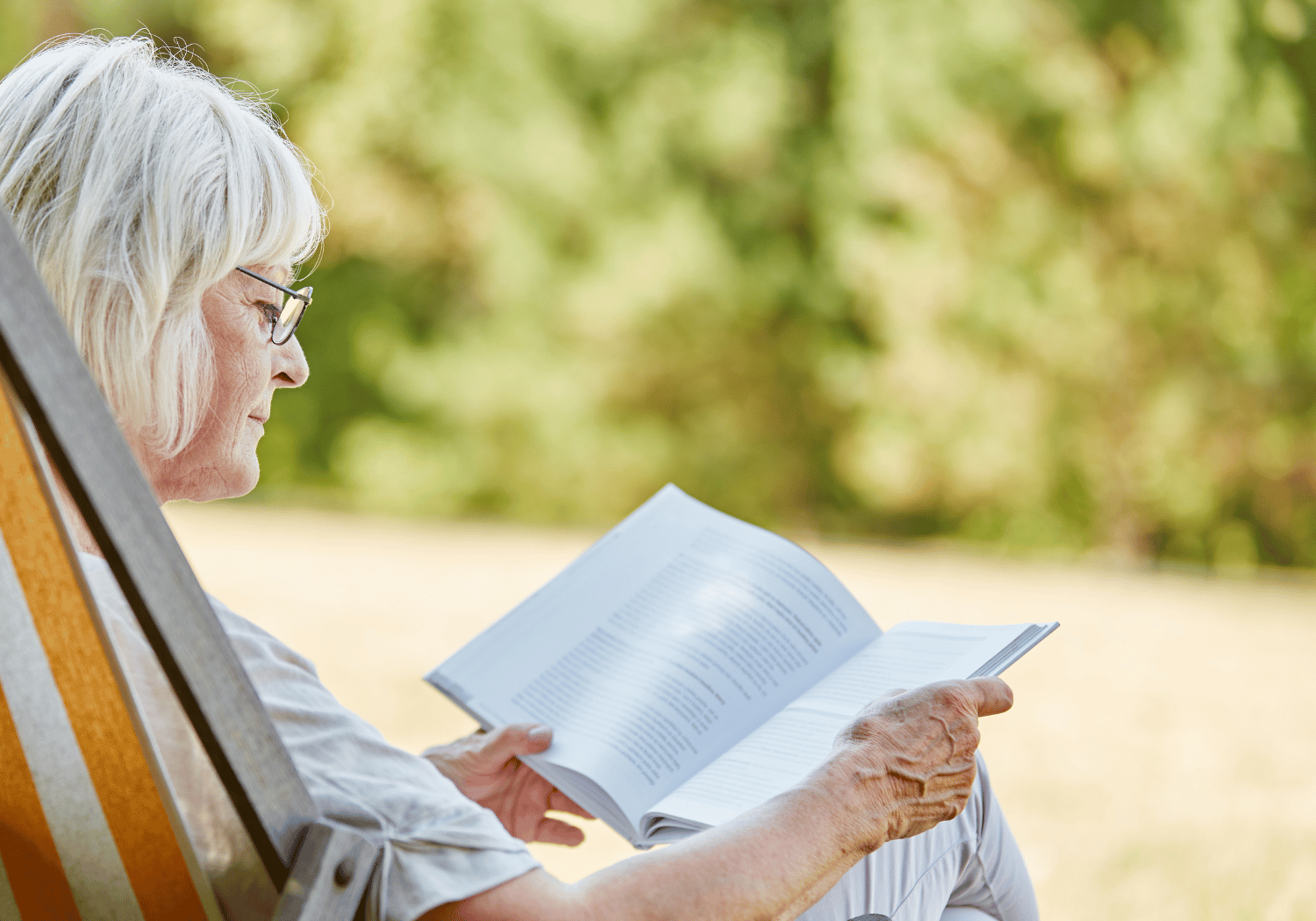 Older woman with white hair and glasses reading a book while sitting in a wooden chair outdoors, sunlight and greenery in the background.